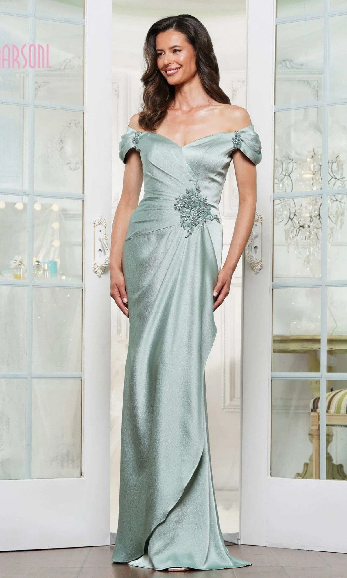 A woman wearing the Marsoni MV1351 long formal dress in sage stands indoors by glass doors. This elegant, off-the-shoulder chiffon gown features floral embellishments at the waist and exudes timeless sophistication.